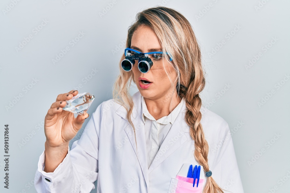 Sticker Beautiful young blonde woman holding brilliant diamond stone wearing magnifier glasses afraid and shocked with surprise and amazed expression, fear and excited face.