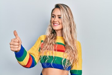 Beautiful young blonde woman wearing colored sweater looking proud, smiling doing thumbs up gesture to the side