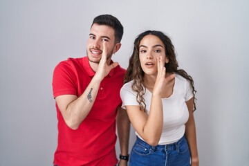 Naklejka premium Young hispanic couple standing over isolated background hand on mouth telling secret rumor, whispering malicious talk conversation