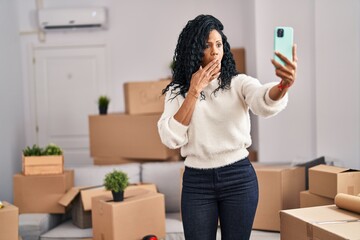 Middle age hispanic woman moving to a new home taking selfie picture covering mouth with hand,...