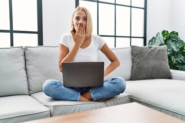 Young caucasian woman using laptop at home sitting on the sofa covering mouth with hand, shocked...