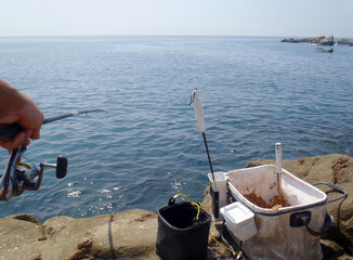 Sunny weather, rocky shore fishing scene. Hand holding rod, ground bait bucket and tackles set on...
