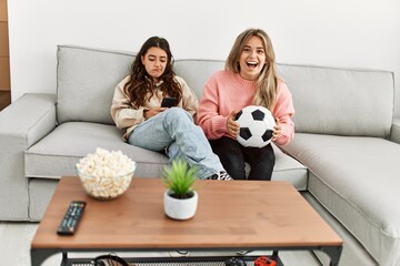 Girl smiling happy watching soccer match and her girlfriend boring using smartphone at home.