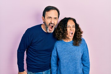 Middle age couple of hispanic woman and man hugging and standing together angry and mad screaming frustrated and furious, shouting with anger. rage and aggressive concept.