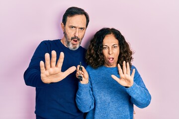 Middle age couple of hispanic woman and man holding keys of new home doing stop gesture with hands palms, angry and frustration expression