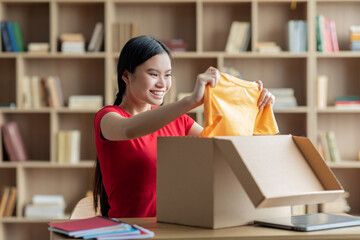 Cheerful young korean woman takes out clothes from cardboard box in living room interior