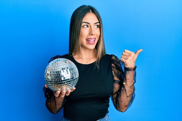 Young hispanic woman holding shiny disco ball pointing thumb up to the side smiling happy with open...