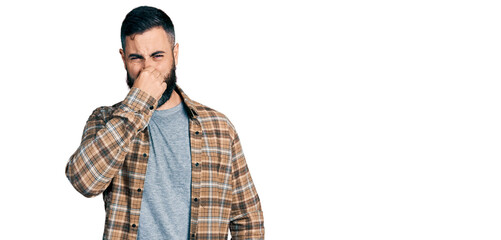 Hispanic man with beard wearing casual shirt smelling something stinky and disgusting, intolerable...