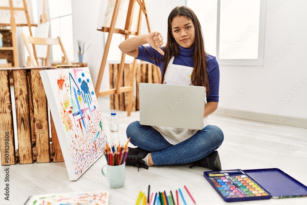 Canvas Prints Young brunette woman sitting on the floor at art studio with laptop with angry face, negative sign showing dislike with thumbs down, rejection concept