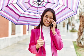 Young latin woman talking on the smartphone holding umbrella at street
