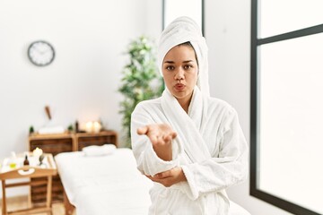 Young brunette woman wearing towel and bathrobe standing at beauty center looking at the camera blowing a kiss with hand on air being lovely and sexy. love expression.