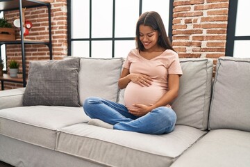 Young latin woman pregnant touching belly sitting on sofa at home