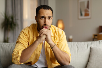 Black man sitting on couch with thoughtful face expression, feeling sad, having problem, suffering...