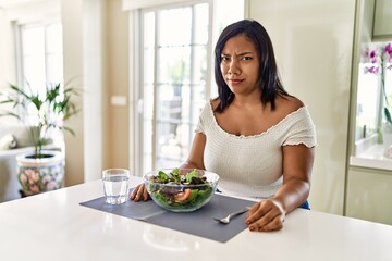 Obraz na płótnie Canvas Young hispanic woman eating healthy salad at home skeptic and nervous, frowning upset because of problem. negative person.