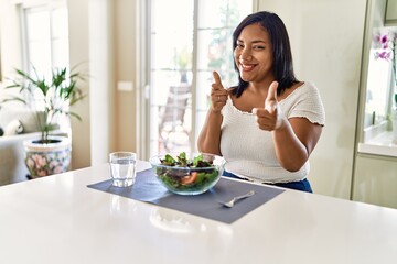 Young hispanic woman eating healthy salad at home pointing fingers to camera with happy and funny...