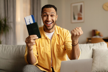 Cool young black man with passport and plane ticket gesturing YES, using laptop to buy tour at travel agency from home - obrazy, fototapety, plakaty