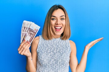 Young caucasian blonde woman holding egyptian pounds banknotes celebrating achievement with happy smile and winner expression with raised hand - obrazy, fototapety, plakaty
