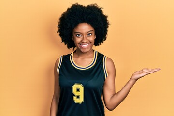 Fototapeta na wymiar Young african american woman wearing basketball uniform smiling cheerful presenting and pointing with palm of hand looking at the camera.