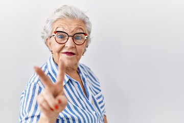 Senior woman with grey hair standing over white background smiling looking to the camera showing fingers doing victory sign. number two.