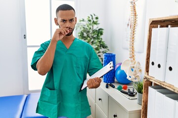 African american physiotherapist man working at pain recovery clinic mouth and lips shut as zip...
