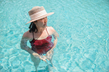 Happy asian woman in red swimsuit and a straw hat relaxing in swimming pool looking at view by the pool at Koh Mak, Phangnga, Thailand. relax and travel concept. Comfort resort summer vacation