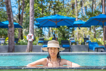 Happy asian woman in red swimsuit and a straw hat relaxing in swimming pool looking at camera by the pool at Koh Mak, Phangnga, Thailand. relax and travel concept. Comfort resort summer vacation