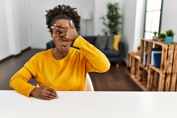 Young african american woman wearing casual clothes sitting on the table at home peeking in shock covering face and eyes with hand, looking through fingers with embarrassed expression.