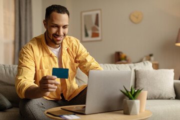 Happy young black guy booking hotel online, using laptop and credit card at home, copy space