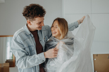 Cheerful young couple in love in their new apartment, having fun with wrapping foil. Conception of moving.