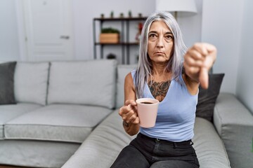 Middle age grey-haired woman drinking coffee sitting on the sofa at home looking unhappy and angry...