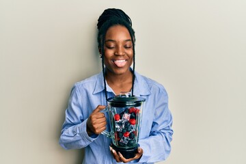 African american woman with braided hair holding food processor mixer machine with fruits sticking tongue out happy with funny expression.
