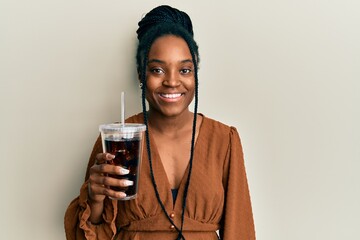 African american woman with braided hair drinking glass of cola beverage looking positive and happy...