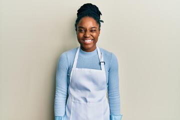 African american woman with braided hair wearing cleaner apron and gloves winking looking at the...