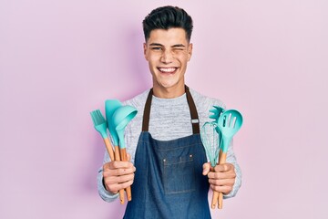 Young hispanic man wearing professional baker apron holding cooking tools winking looking at the...