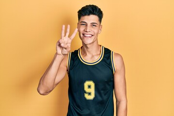 Young hispanic man wearing basketball uniform showing and pointing up with fingers number three...