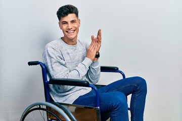 Young hispanic man sitting on wheelchair clapping and applauding happy and joyful, smiling proud hands together