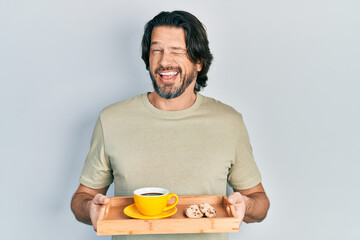 Middle age caucasian man holding breakfast tray with cookies and coffee winking looking at the camera with sexy expression, cheerful and happy face.