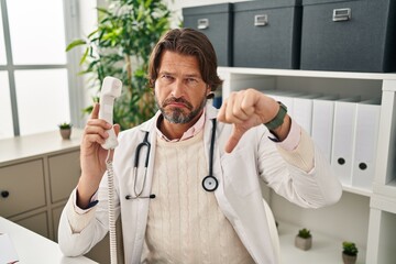 Handsome middle age doctor man speaking on the phone at the clinic with angry face, negative sign showing dislike with thumbs down, rejection concept