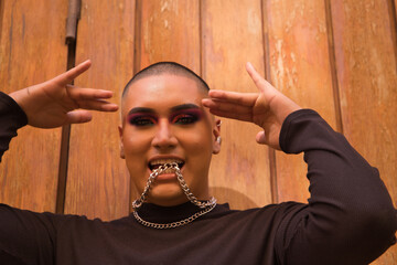 Portrait of non-binary person, young and South American, heavily make up biting a metal chain with teeth, with a wooden wall in the background. Concept queen, lgbtq+, pride, queer.