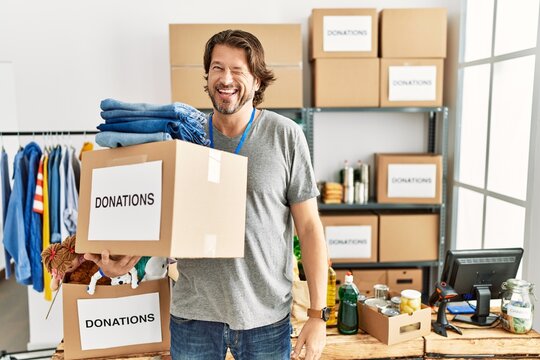Handsome middle age man holding donations box for charity at volunteer stand winking looking at the camera with sexy expression, cheerful and happy face.