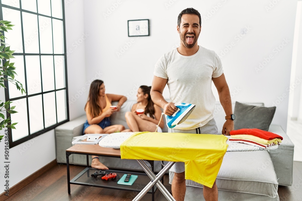 Wall mural young hispanic man ironing clothes at home sticking tongue out happy with funny expression. emotion 