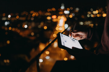 Close-up of man standing on balcony with urban view and using tablet at night