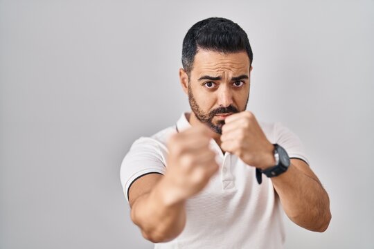Young hispanic man with beard wearing casual clothes over white background ready to fight with fist defense gesture, angry and upset face, afraid of problem