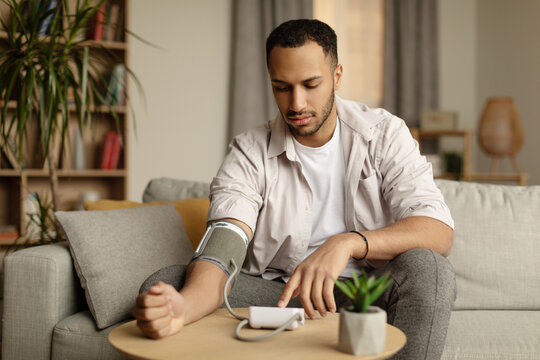 Young Black Man Sitting On Couch, Checking Blood Pressure At Home, Copy Space