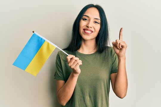 Young hispanic girl wearing ukraine flag smiling with an idea or question pointing finger with happy face, number one