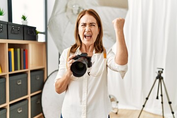 Middle age photography woman using camera at photo studio annoyed and frustrated shouting with anger, yelling crazy with anger and hand raised