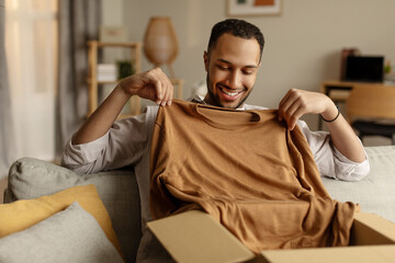 Cheerful black guy unboxing cardboard parcel, satisfied with great purchase, taking out new clothes...