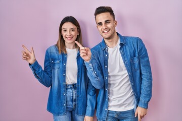 Young hispanic couple standing over pink background with a big smile on face, pointing with hand finger to the side looking at the camera.