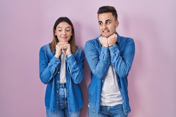 Young hispanic couple standing over pink background laughing nervous and excited with hands on chin...