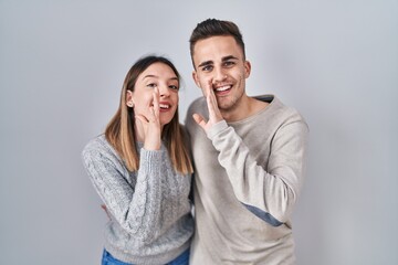 Young hispanic couple standing over white background hand on mouth telling secret rumor, whispering...
